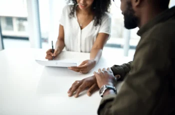 Duas pessoas analisam e assinam um documento sobre uma mesa em um ambiente de escritório.