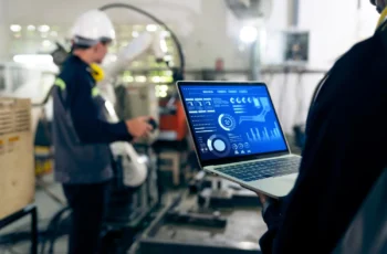 Um técnico em ambiente industrial utiliza um laptop com painel digital enquanto outro trabalhador opera equipamentos ao fundo.