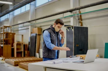 Profissional em ambiente industrial analisando projetos sobre uma mesa, com notebook ao lado e estruturas de móveis ao fundo.